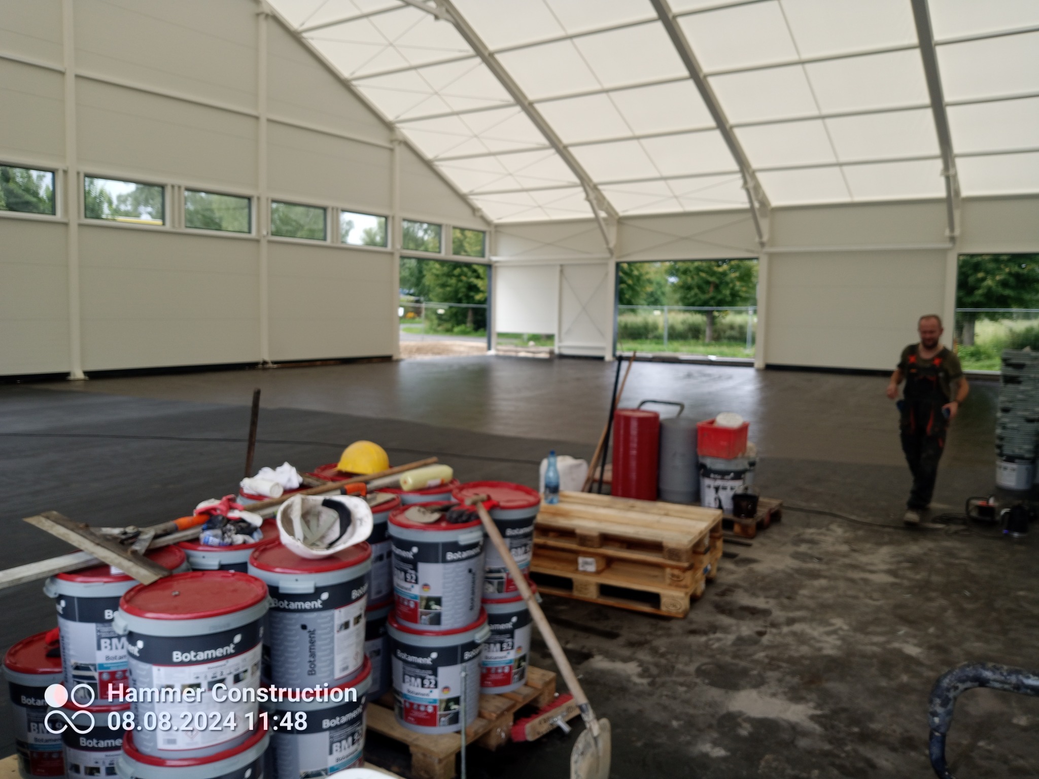 Wnętrze hali z przygotowaną, świeżo wylaną posadzką, widoczne wiadra z materiałami Botament BM 92, palety, narzędzia i pracownik w tle, wpadające światło przez okna.