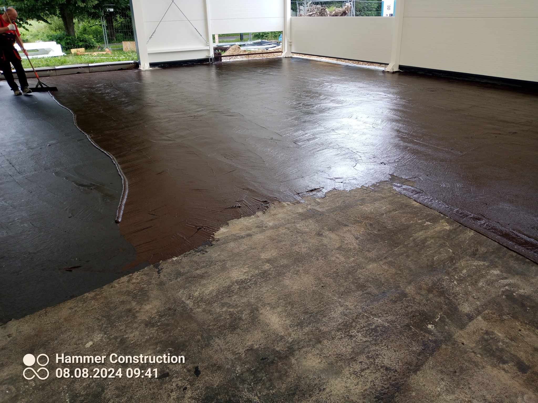 Aplikacja brązowej powłoki na betonowej posadzce wewnątrz hali, widoczny pracownik rozprowadzający materiał za pomocą rakli.