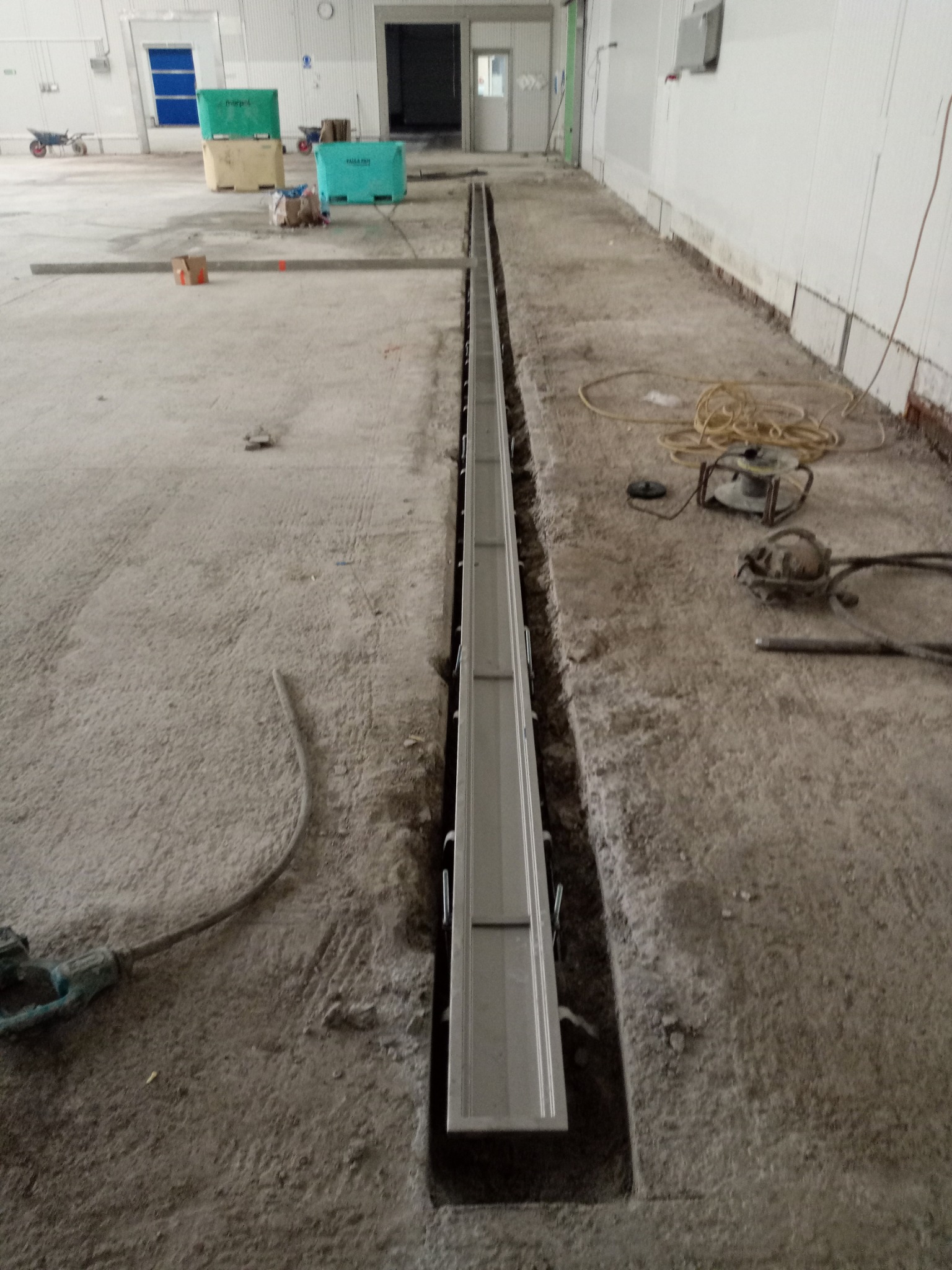 Montaż liniowego odwodnienia podłogowego w hali przemysłowej, widoczne narzędzia budowlane i fragmenty posadzki przed wylewką.