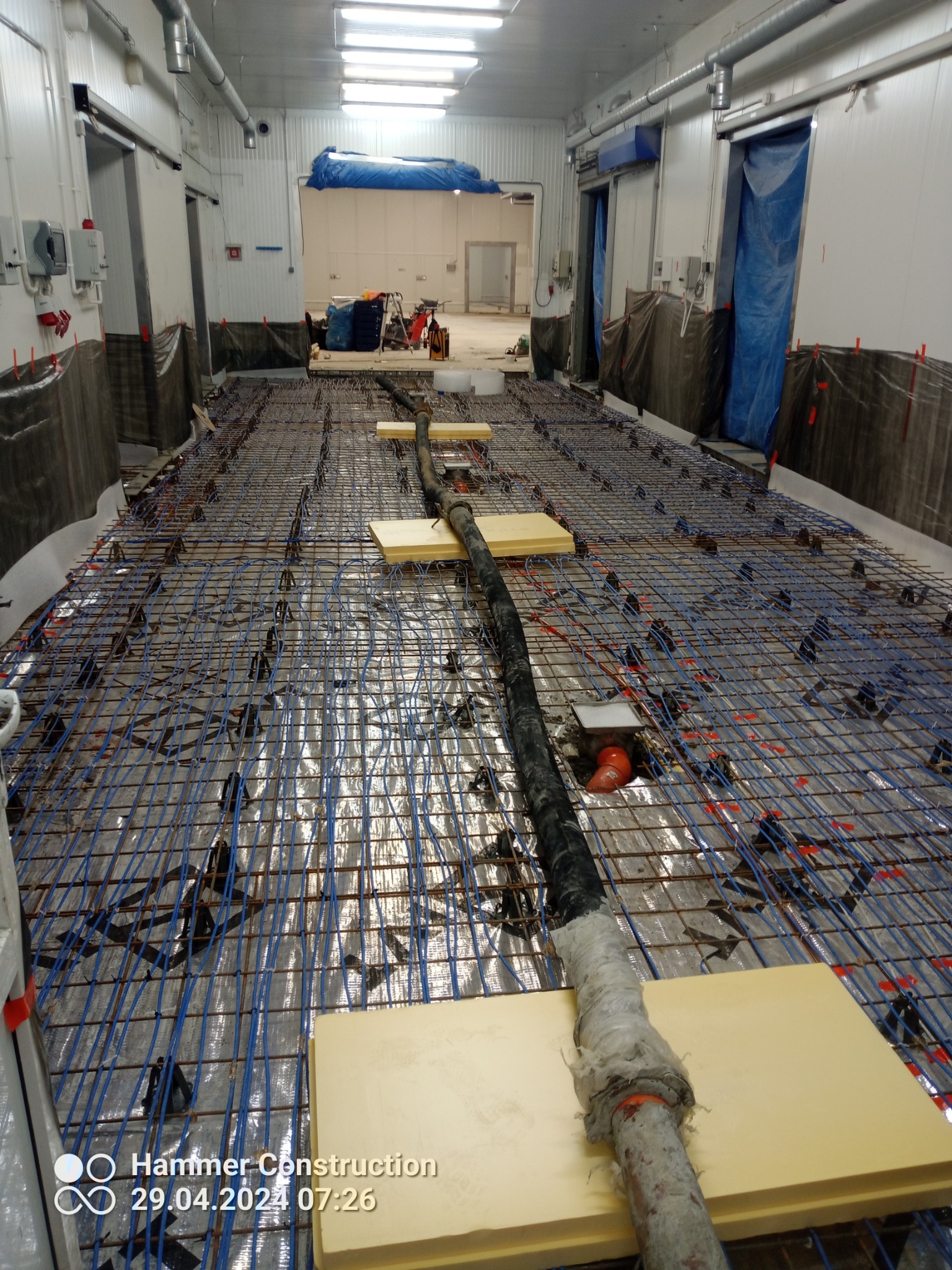 Przygotowanie do wylewki w hali przemysłowej: siatka zbrojeniowa, ogrzewanie podłogowe i rury na izolacji, widoczne płyty styropianowe i folia ochronna.