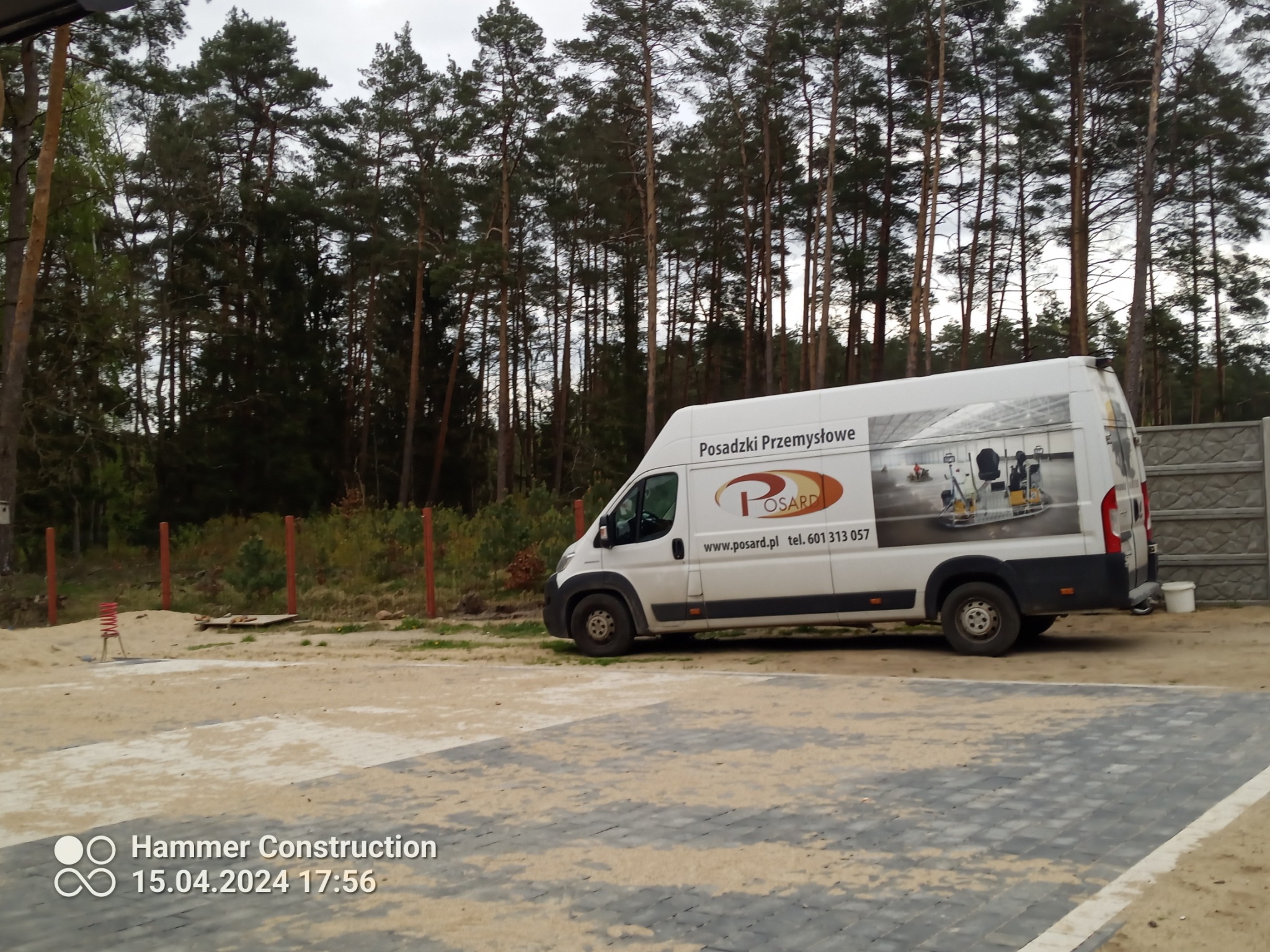 Biały samochód dostawczy z logo firmy i napisem 'Posadzki Przemysłowe' zaparkowany na placu z kostką brukową, w tle las i ogrodzenie.