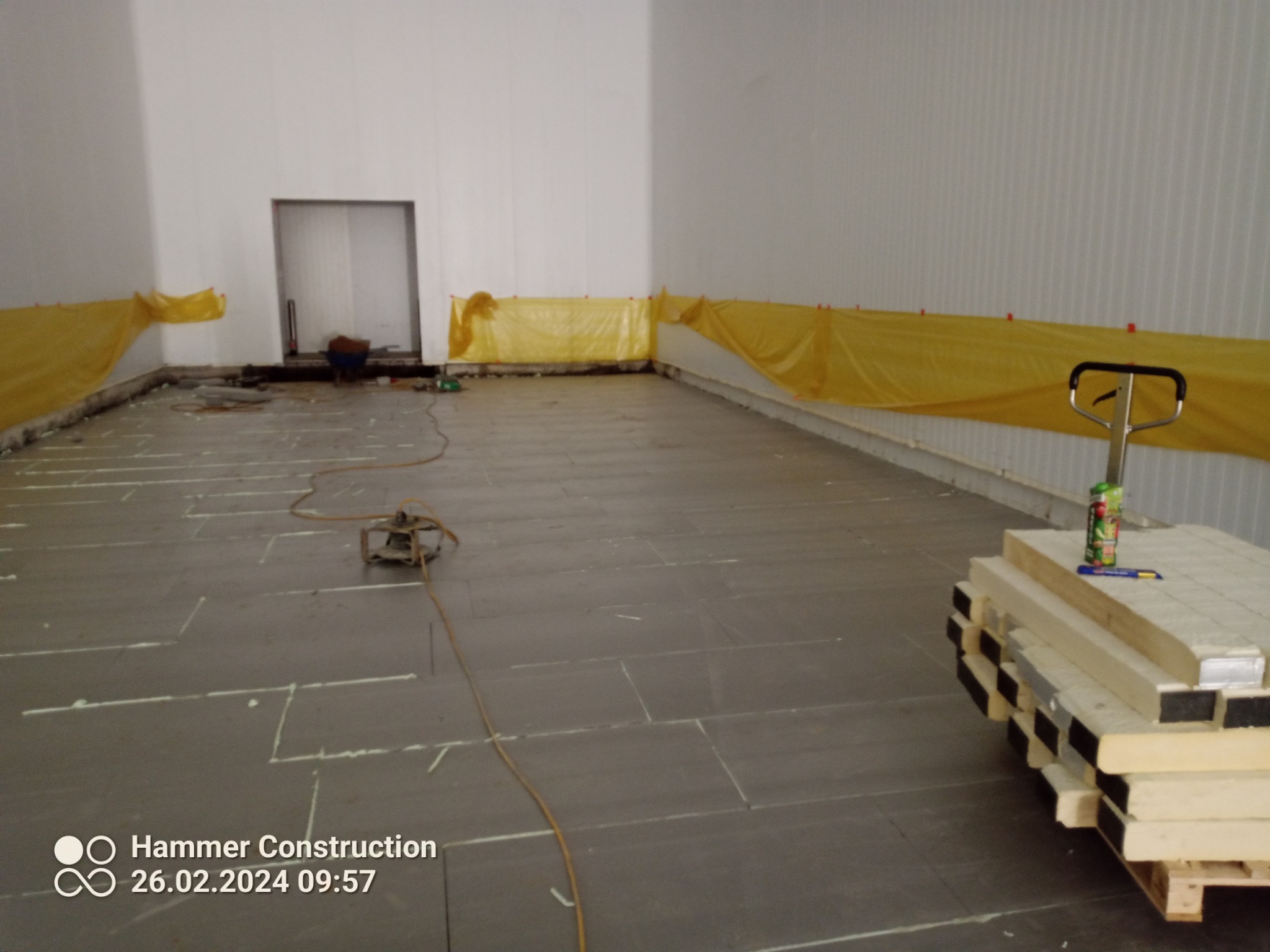 Przygotowanie podłoża w hali produkcyjnej: rozłożone płyty izolacyjne z widocznymi liniami cięcia, przykryte folią ochronną ściany, oraz stos płyt na palecie z narzędziami i napojem energetycznym.