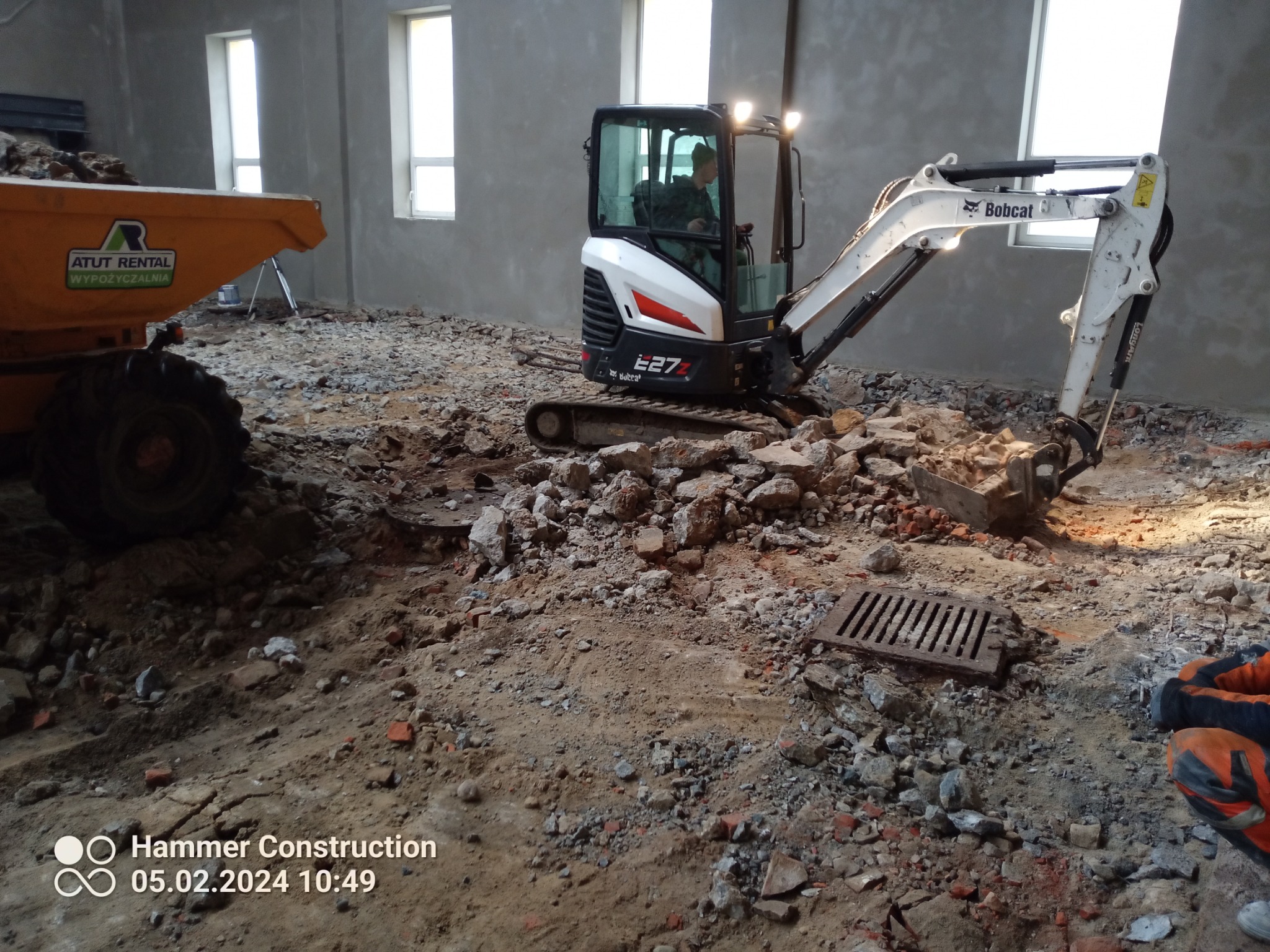 Mini koparka Bobcat E27Z w trakcie prac przygotowawczych do wylewki wewnątrz budynku, widoczny operator, gruz, piasek i częściowo załadowana wywrotka ATUT Rental w tle.