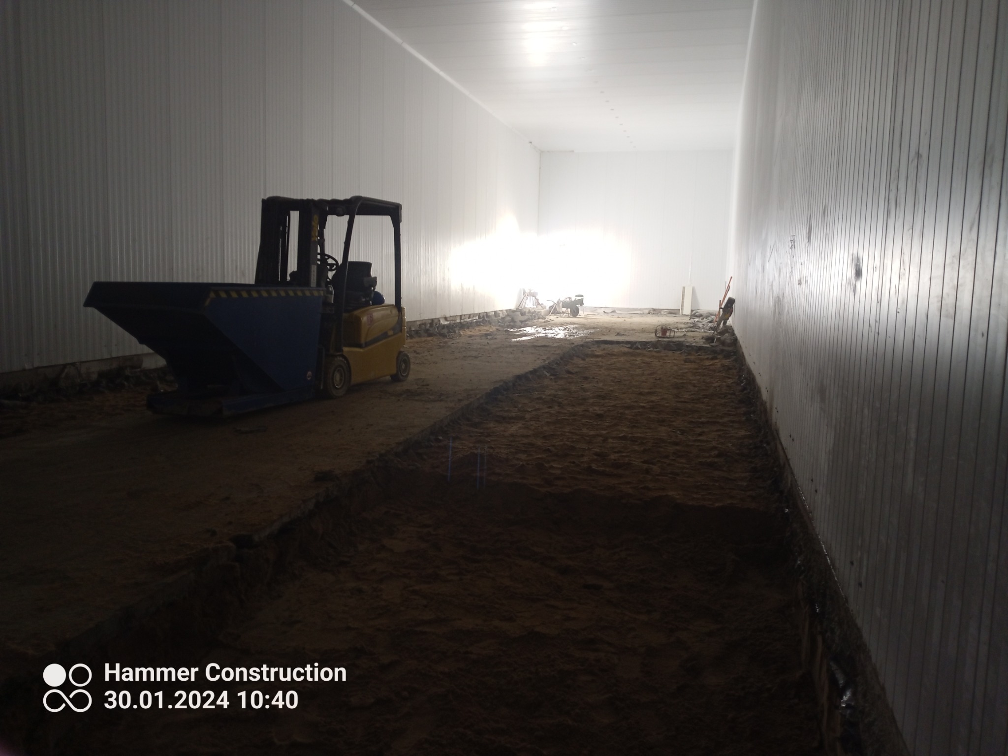 Wnętrze hali w trakcie przygotowania podłoża, widoczny wózek widłowy z niebieskim pojemnikiem, ziemia wyrównana na różnych poziomach, białe panele ścienne.