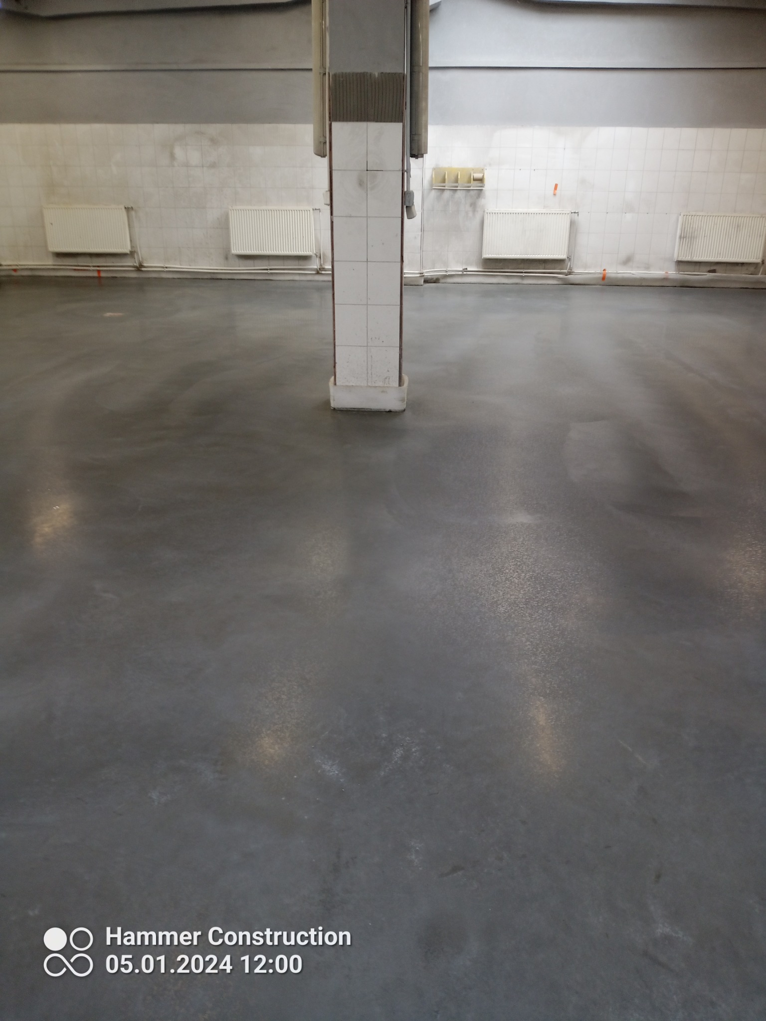 Szara, błyszcząca posadzka w hali przemysłowej z białymi płytkami na ścianach i grzejnikami, widoczny słup podtrzymujący strop.