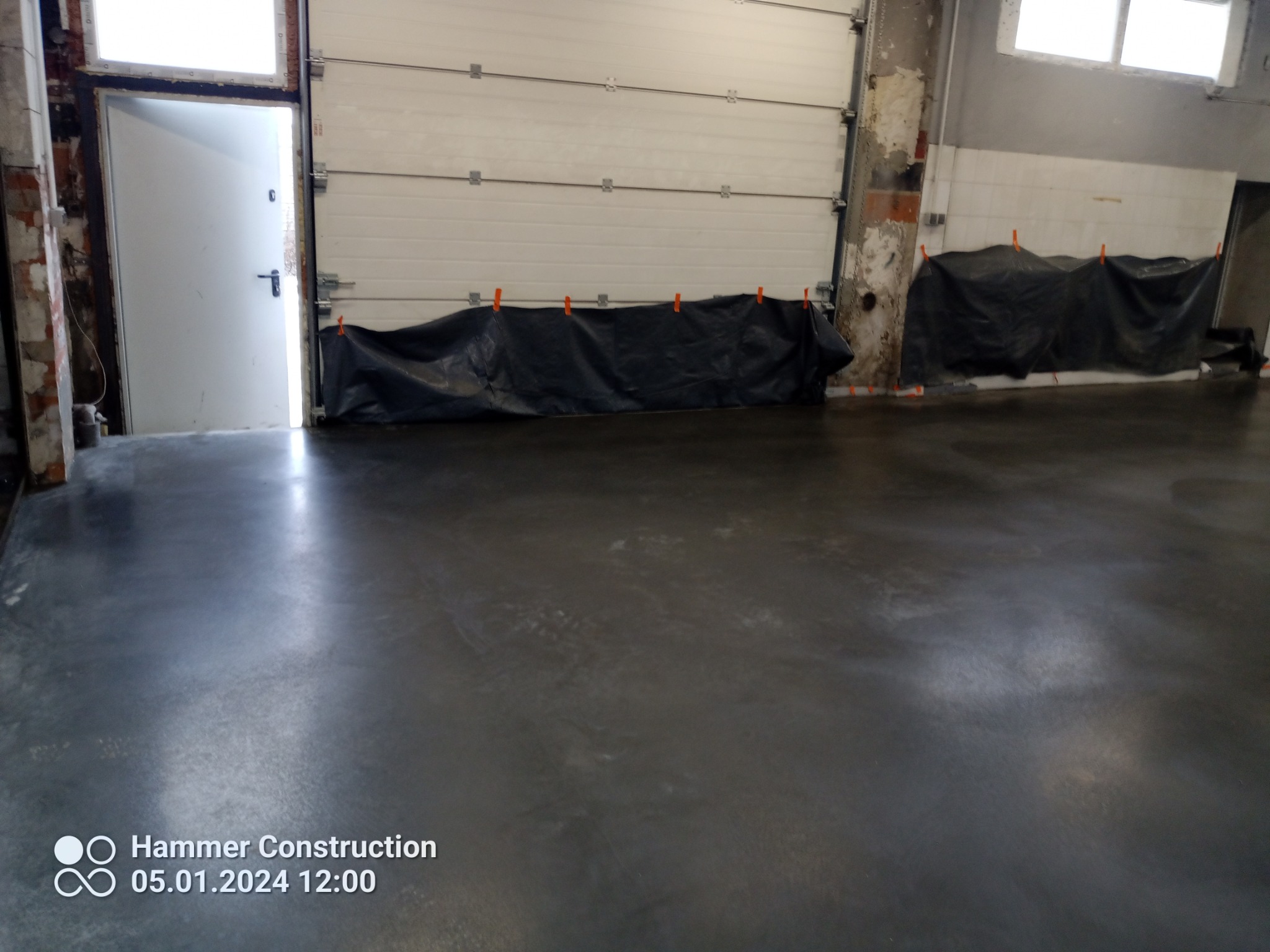 Świeżo wylana, szara posadzka przemysłowa w hali, zabezpieczona folią przy bramie garażowej i ścianach, widoczne ślady po procesie wylewania.