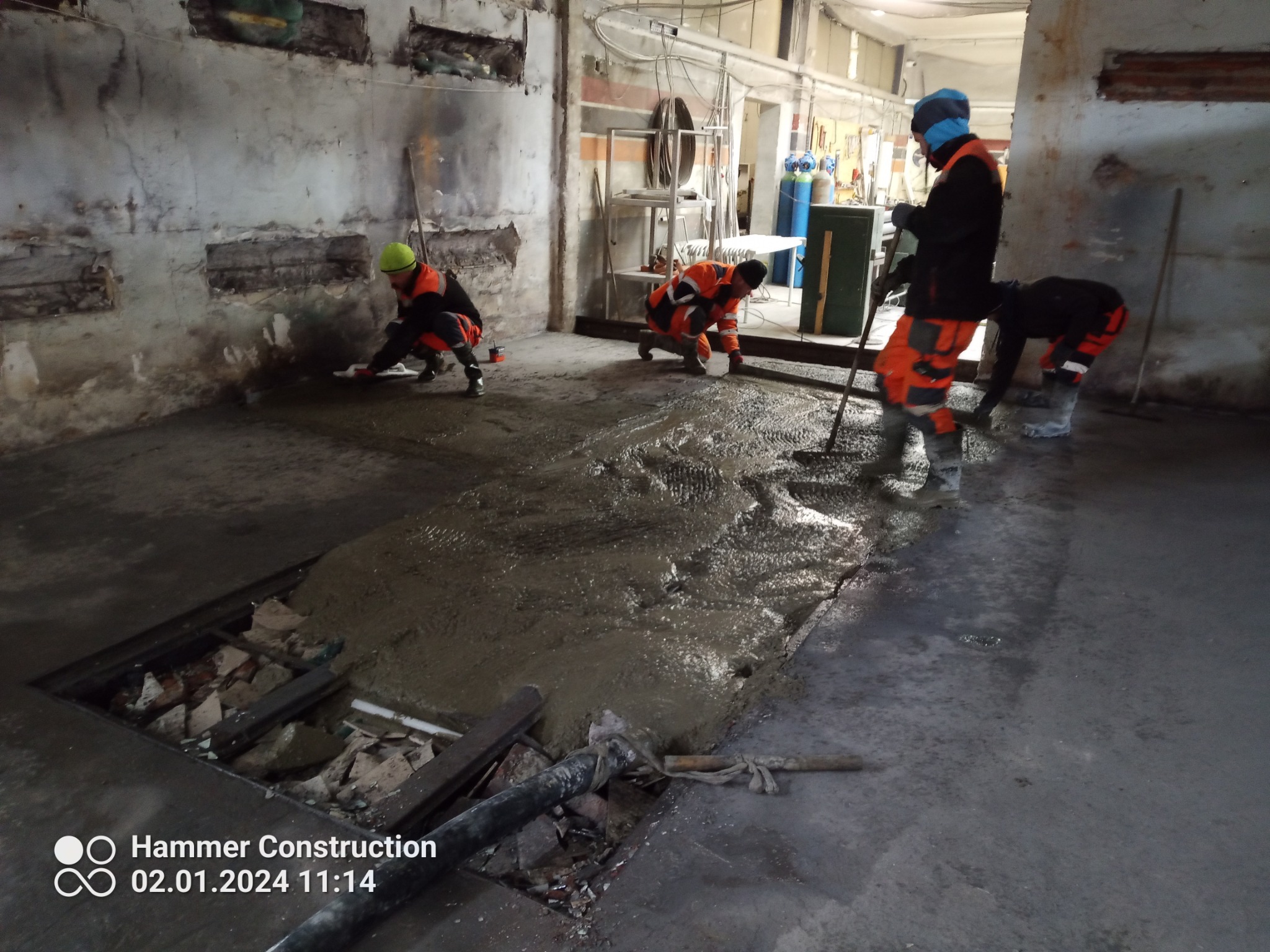 Robotnicy w pomarańczowych kombinezonach wyrównują szarą, mokrą wylewkę w surowym, przemysłowym wnętrzu z odsłoniętą cegłą i instalacjami.