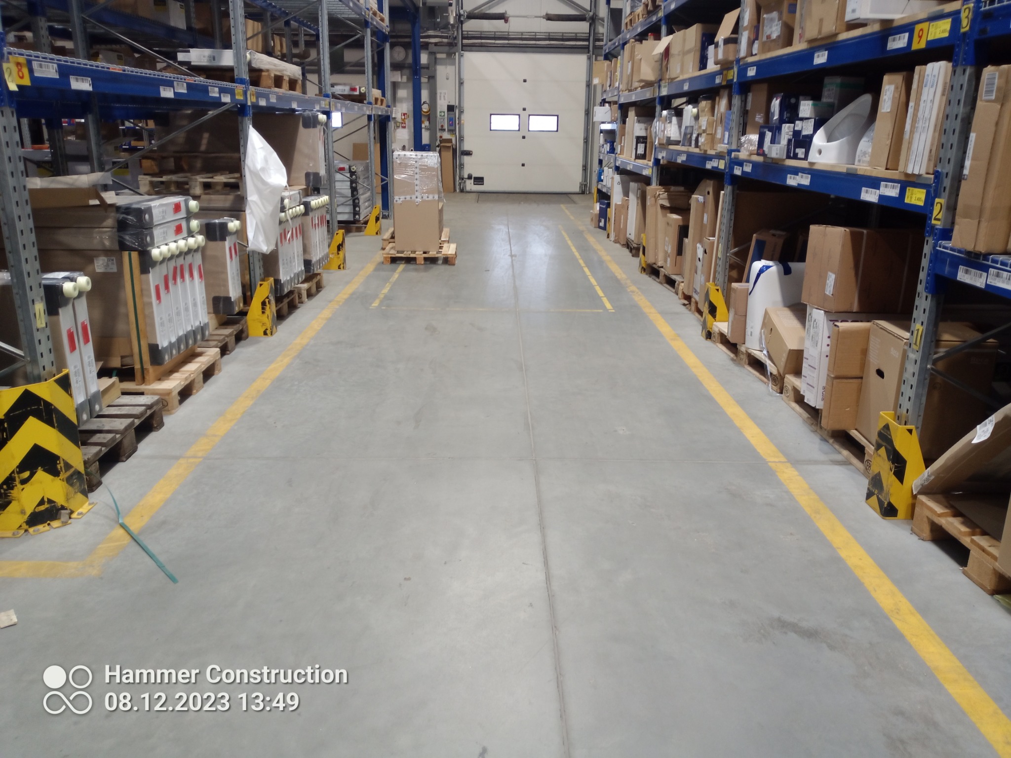 Magazyn z szarą, betonową posadzką, wyznaczonymi żółtymi liniami transportowymi i regałami wypełnionymi kartonami po bokach; w tle brama wjazdowa.