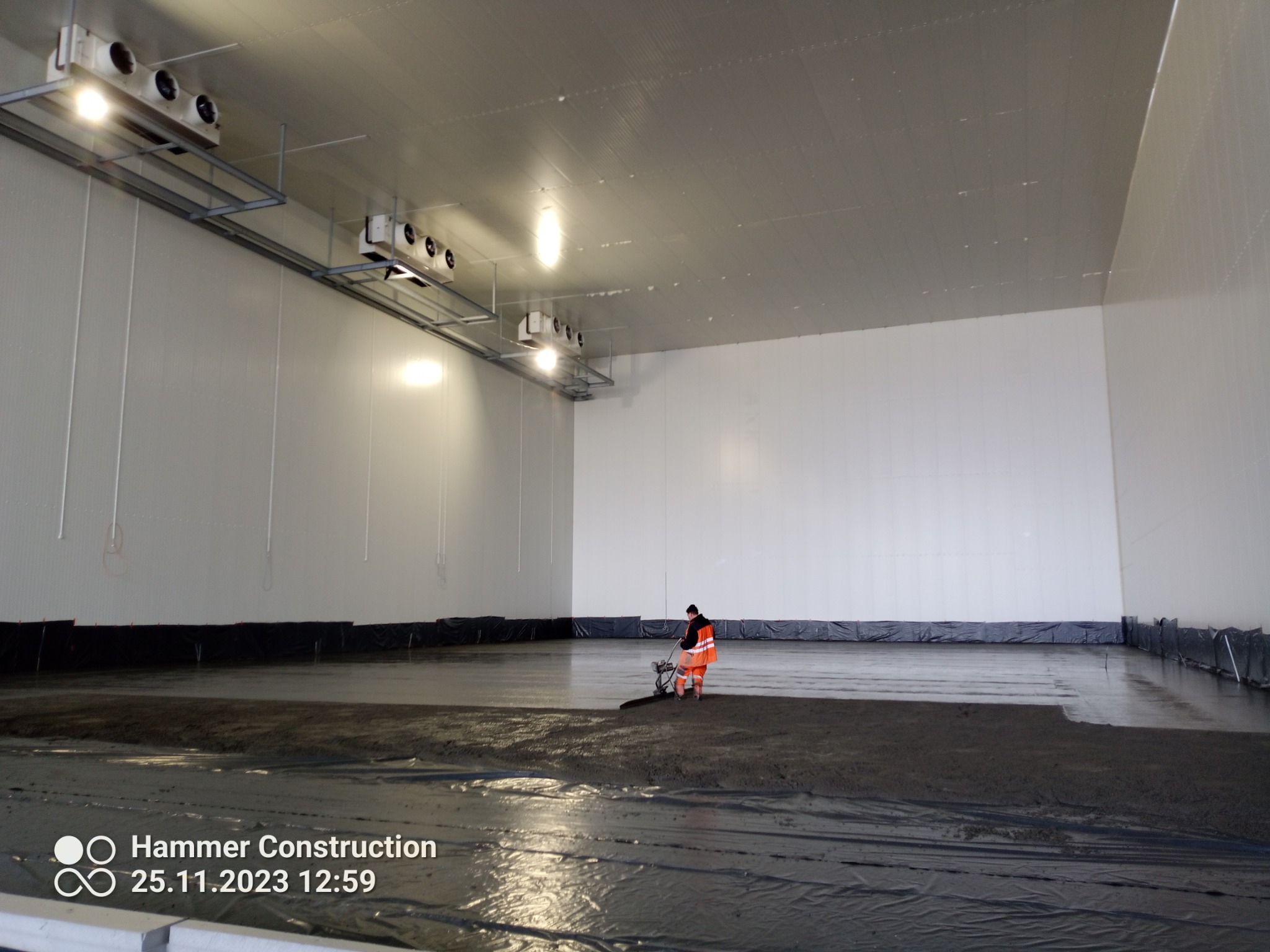 Pracownik w odblaskowej kamizelce wyrównuje masę posadzkową w przemysłowym wnętrzu hali, widoczne czarne folie zabezpieczające ściany, oświetlenie sufitowe i wentylacja.