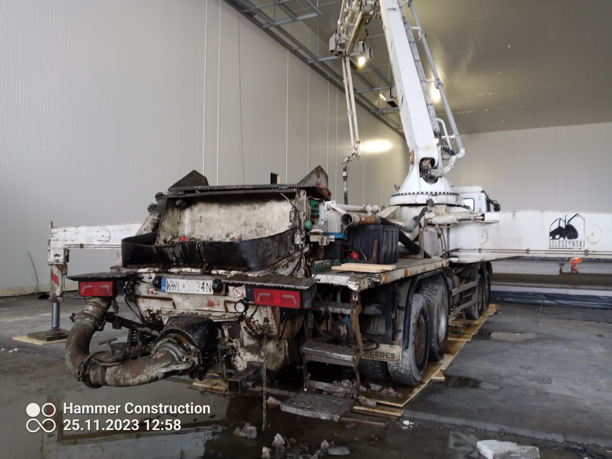 Widok z tyłu brudnej pompy do betonu na podwoziu ciężarówki, zaparkowanej w hali z szarymi ścianami z blachy falistej, z widocznym wysięgnikiem i elementami hydraulicznymi.