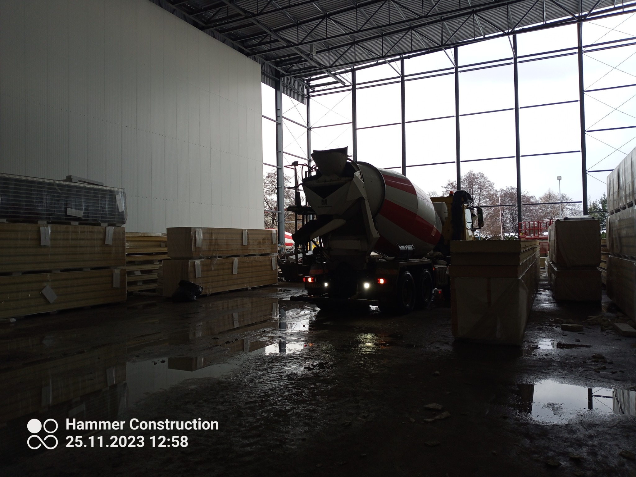 Ciężarówka z betonomieszarką wewnątrz hali przemysłowej, obok składowane płyty warstwowe, mokra posadzka odbija światło.