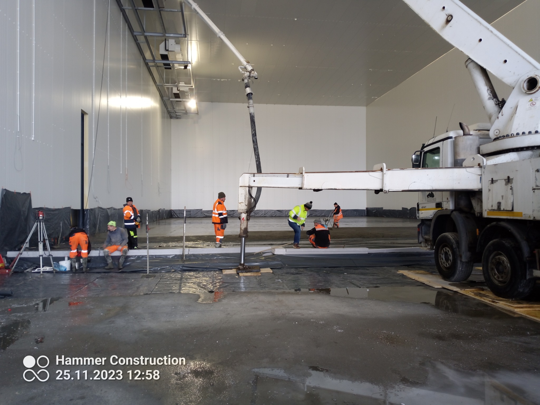 Wnętrze hali z ekipą w strojach roboczych w trakcie wylewania posadzki, widoczna pompa do betonu i rozłożona folia ochronna.
