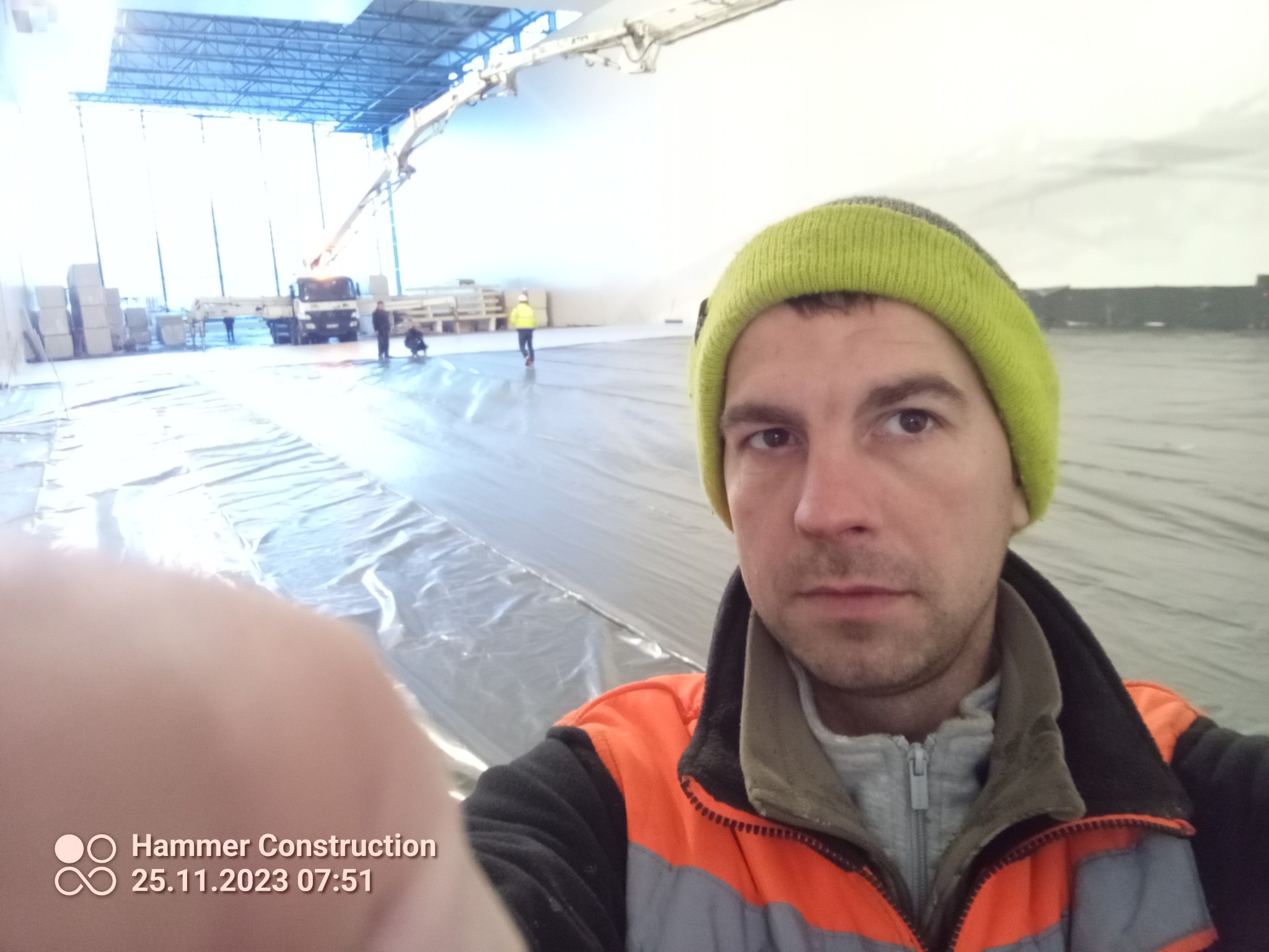Pracownik w czapce i roboczej kurtce robi selfie na tle rozłożonej folii, przygotowanej pod wylewkę anhydrytową w hali przemysłowej, widoczny samochód z pompą do wylewek.