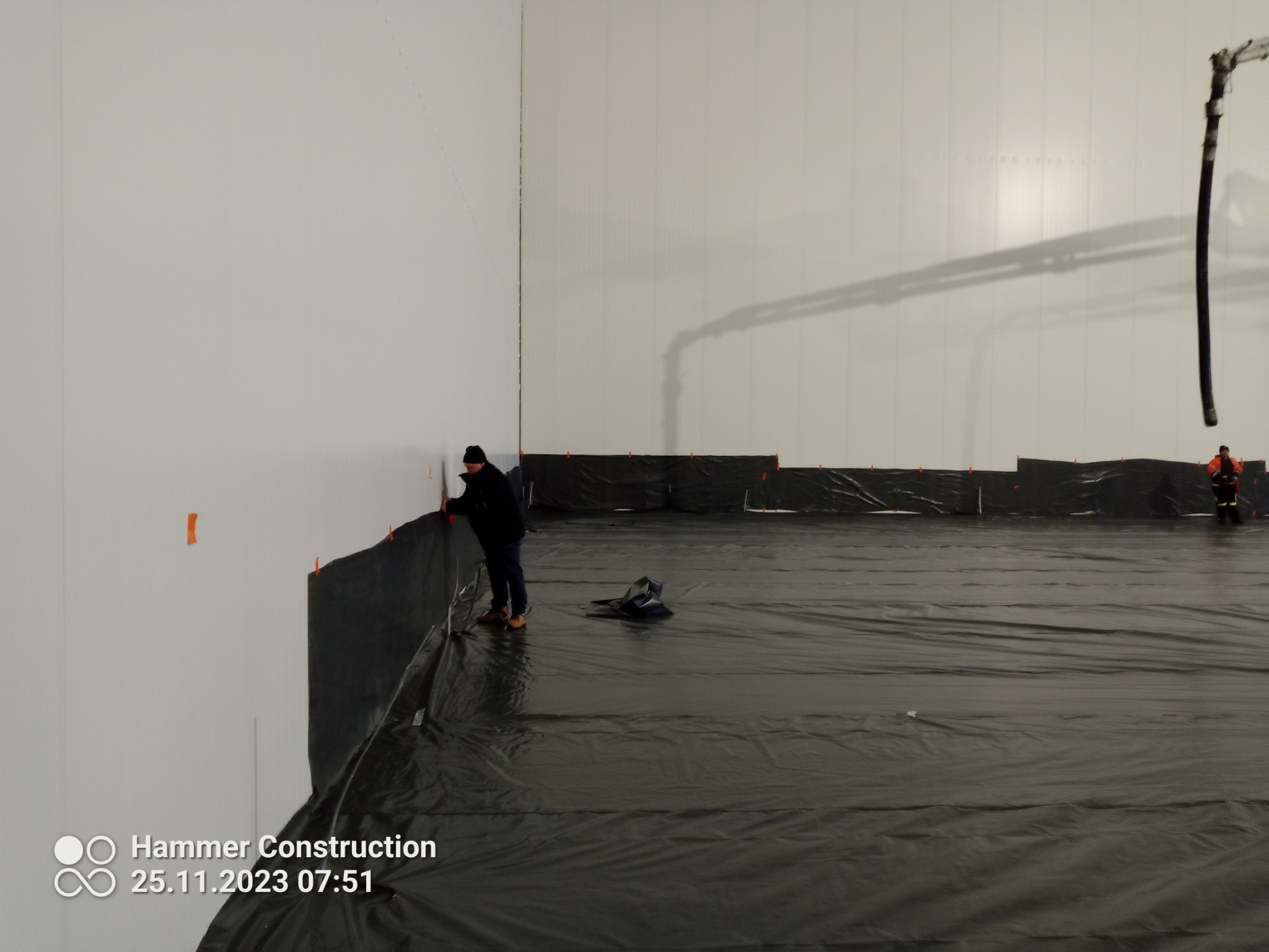 Przygotowanie podłoża w hali przemysłowej: pracownik mocuje folię do ściany, w tle widoczny wąż do wylewki i drugi pracownik.