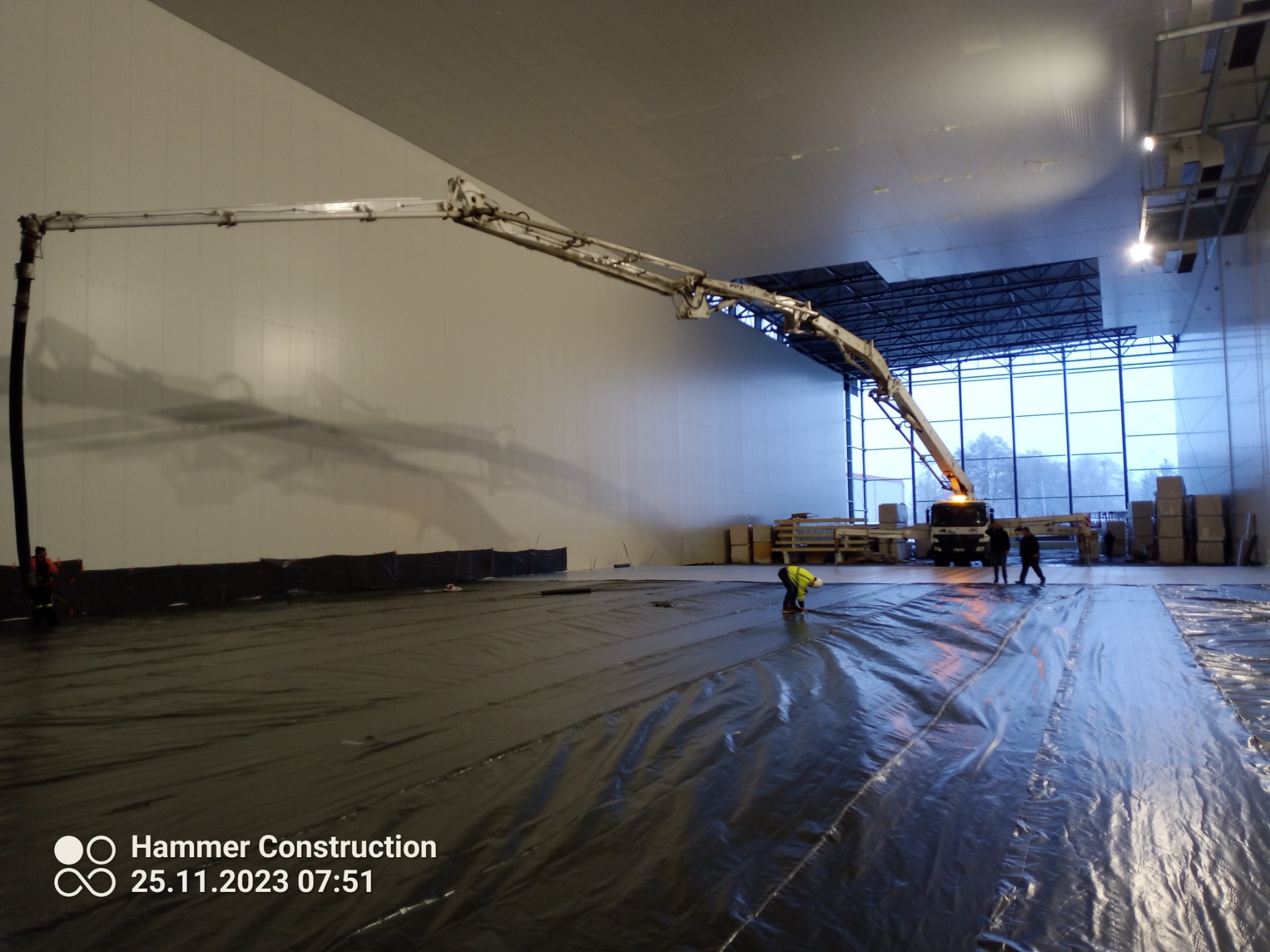 Wnętrze hali przemysłowej z rozłożoną czarną folią, pracownik w żółtej kamizelce wygładza powierzchnię, w tle widoczny samochód z pompą do betonu i grupa osób.