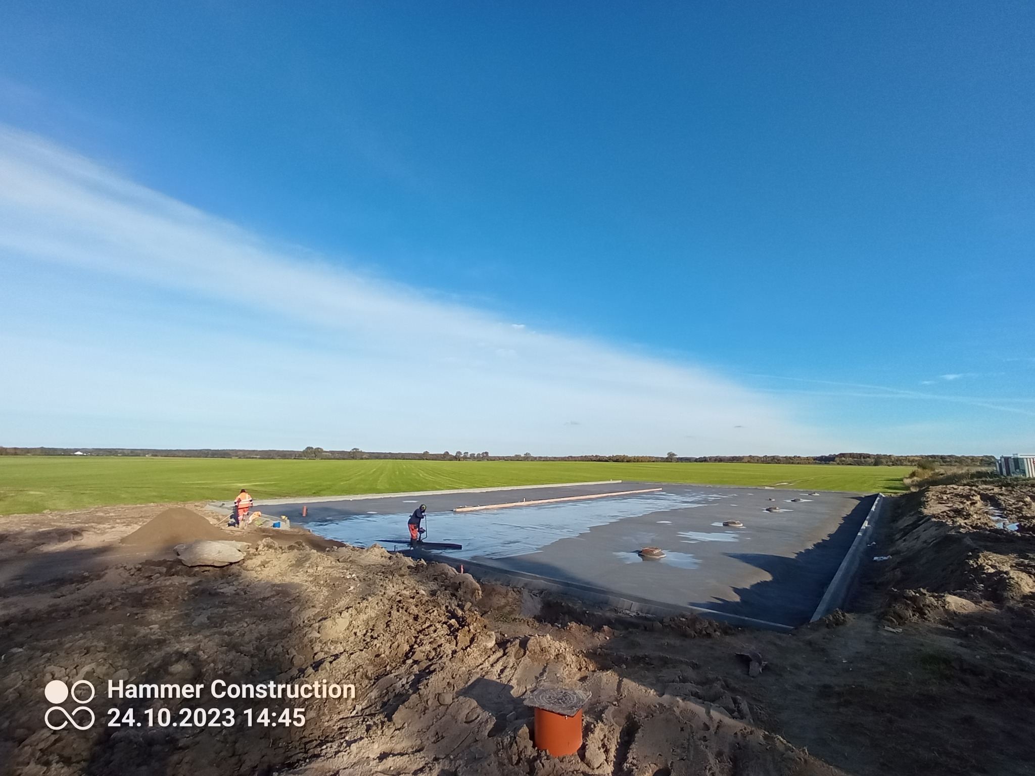 Świeżo wylana, gładka posadzka na placu budowy, dwóch pracowników w odblaskowych ubraniach wyrównuje powierzchnię, w tle zielone pole i błękitne niebo z chmurami.