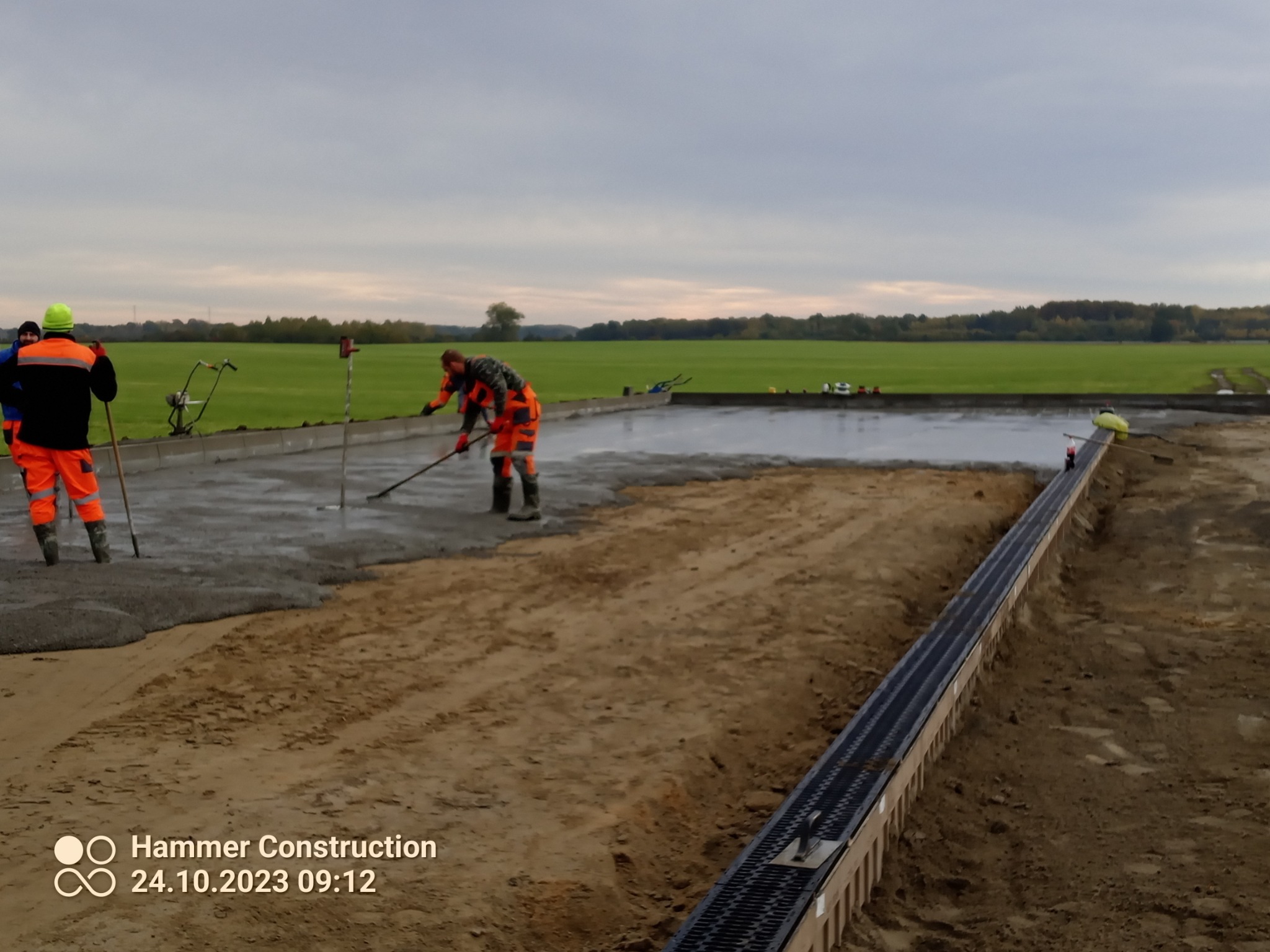 Ekipa budowlana w pomarańczowych kombinezonach rozprowadza świeżą wylewkę na rozległym terenie, z widoczną kratką odwadniającą wzdłuż krawędzi i rozległym zielonym polem w tle pod pochmurnym niebem.