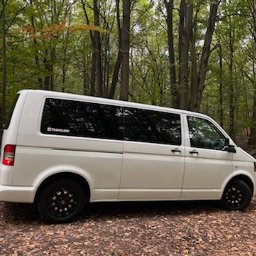 TRAVELSEO MARTA MATERNE - Biały van z czarnymi felgami zaparkowany na leśnej drodze pokrytej liśćmi, z naklejką 'TRAVELAND' na tylnej szybie.
