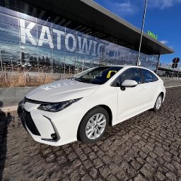 TRAVELSEO MARTA MATERNE - Biała Toyota Corolla zaparkowana na brukowanej nawierzchni przed budynkiem lotniska Katowice-Pyrzowice w słoneczny dzień.