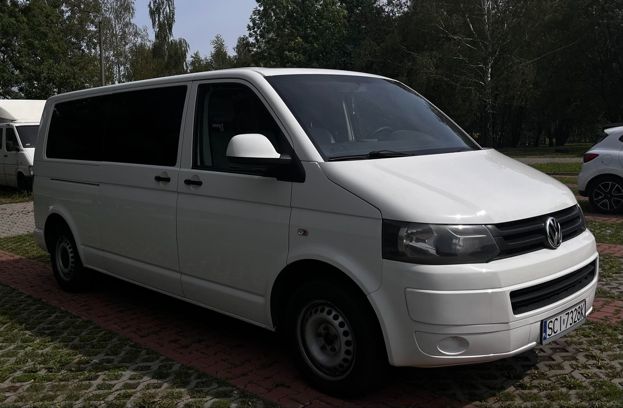 Biały Volkswagen Transporter T5 z przyciemnianymi szybami, zaparkowany na kostce brukowej, widoczny numer rejestracyjny.
