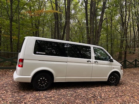 Biały van z czarnymi felgami zaparkowany na leśnej drodze pokrytej liśćmi, z naklejką 'TRAVELAND' na tylnej szybie.
