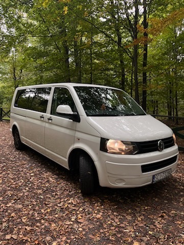 Biały bus Volkswagen Transporter zaparkowany na leśnej drodze pokrytej opadłymi liśćmi, widoczny numer rejestracyjny SC1 72260.