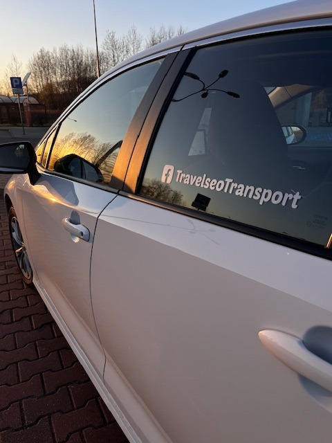Biały samochód osobowy z naklejką 'TravelseoTransport' i logo Facebooka na tylnej szybie, sfotografowany z boku na tle zachodzącego słońca.