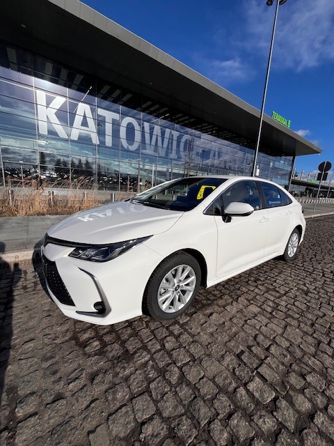 Biała Toyota Corolla zaparkowana na brukowanej nawierzchni przed budynkiem lotniska Katowice-Pyrzowice w słoneczny dzień.