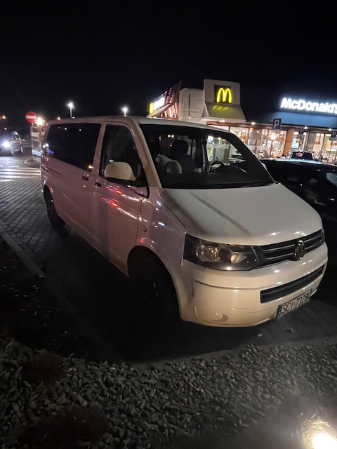 Biały van Volkswagen Transporter zaparkowany nocą na parkingu z widocznym logo McDonald's w tle.