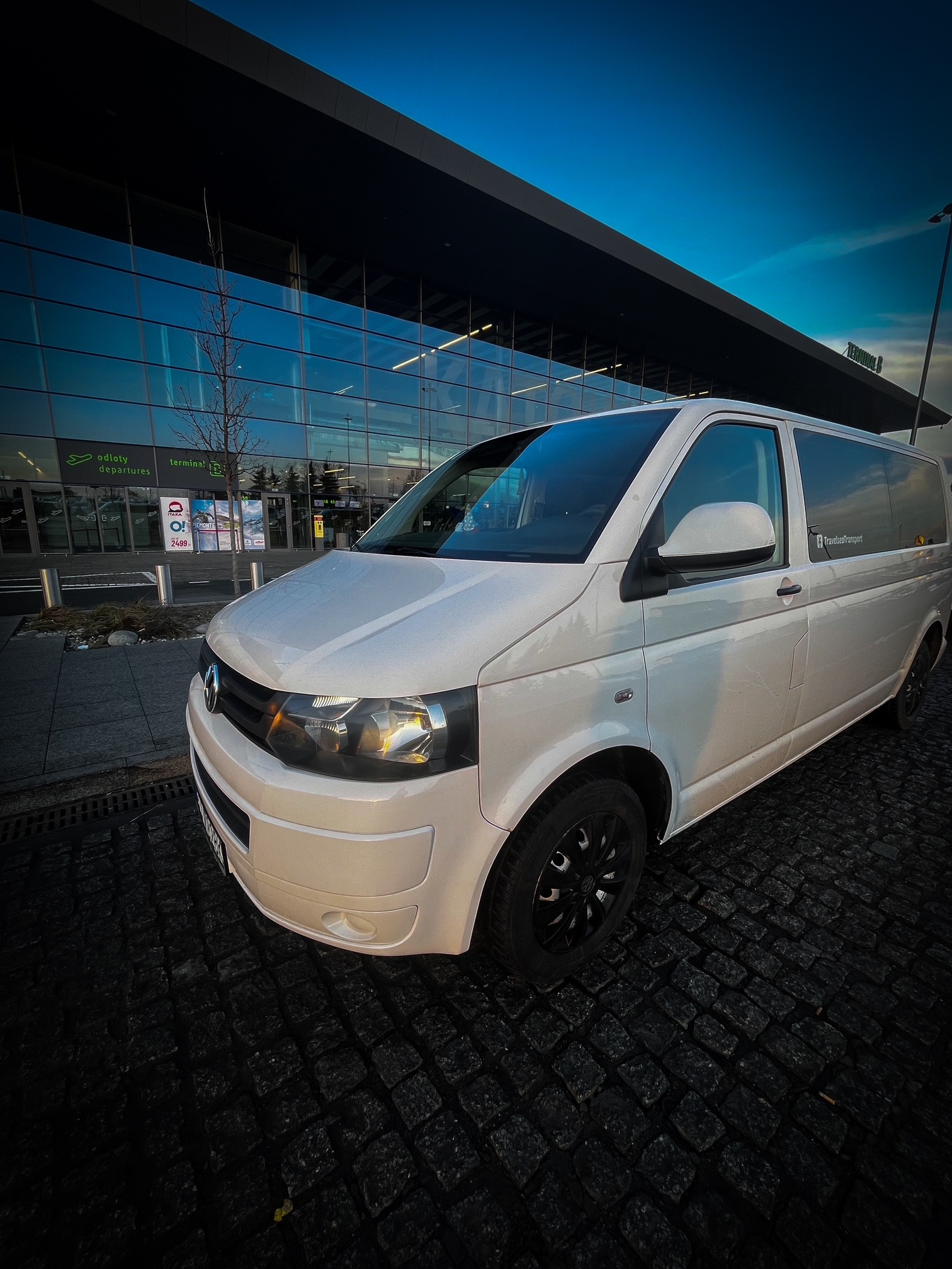 Biały van z czarnymi felgami zaparkowany przed nowoczesnym budynkiem terminalu lotniska, widoczne odbicia w szklanej fasadzie i kostka brukowa na pierwszym planie.