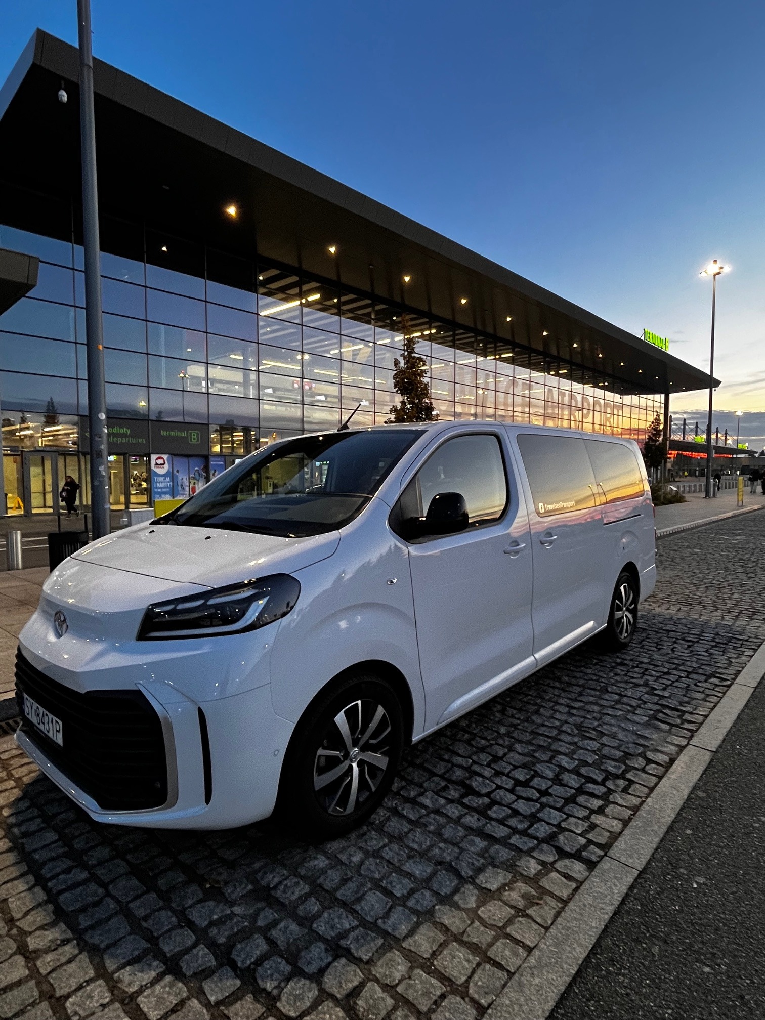 Biały van Toyota Proace Verso na brukowanym parkingu przed lotniskiem o zmierzchu. Widoczne odbicia w szybach i tablica rejestracyjna.