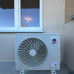 Zewnętrzna jednostka klimatyzacji marki Gree, zamontowana na elewacji budynku z widocznym oknem, oświetlonym żarówką w odbiciu szyby.