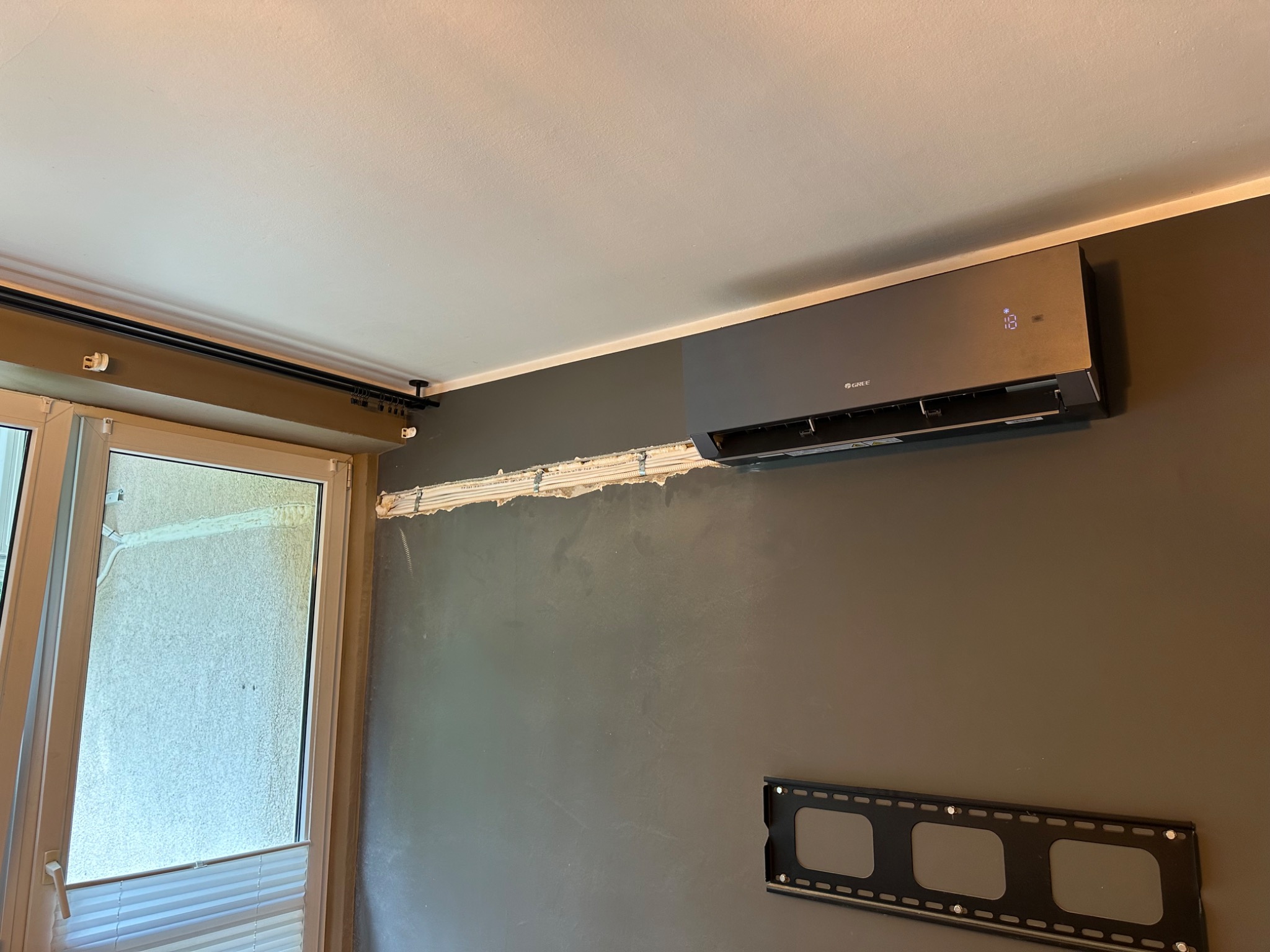 Szary klimatyzator marki Gree zamontowany na szarej ścianie w pomieszczeniu, widoczny uchwyt do telewizora i ślady po montażu instalacji, okno z żaluzjami w tle.