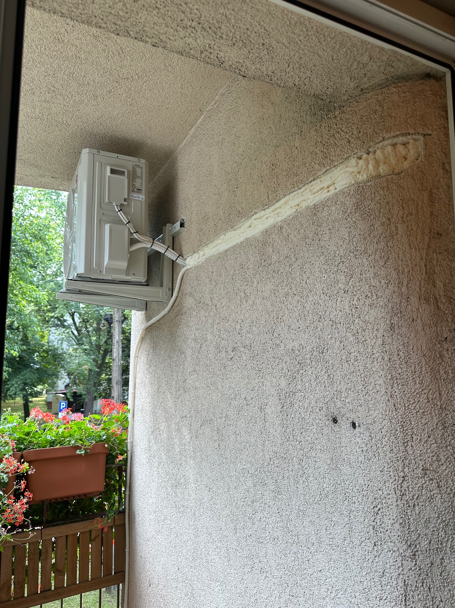 Zewnętrzna jednostka klimatyzacji zamontowana na balkonie z widoczną pianką uszczelniającą wokół rur, kwiaty w doniczce na pierwszym planie.