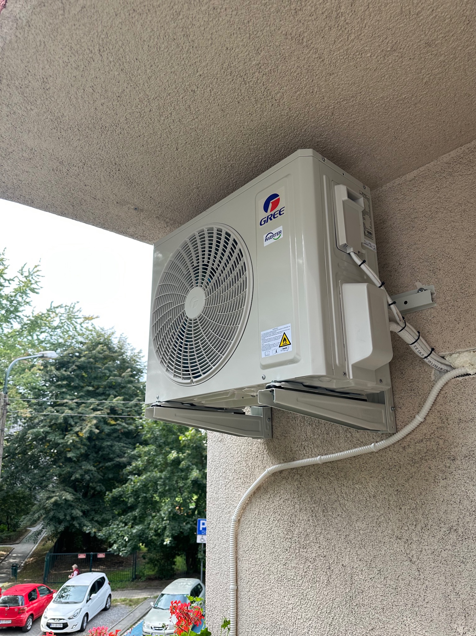 Zewnętrzna jednostka klimatyzacji Gree zamontowana na elewacji budynku, widoczne przewody i uchwyty montażowe, w tle drzewa i zaparkowane samochody.