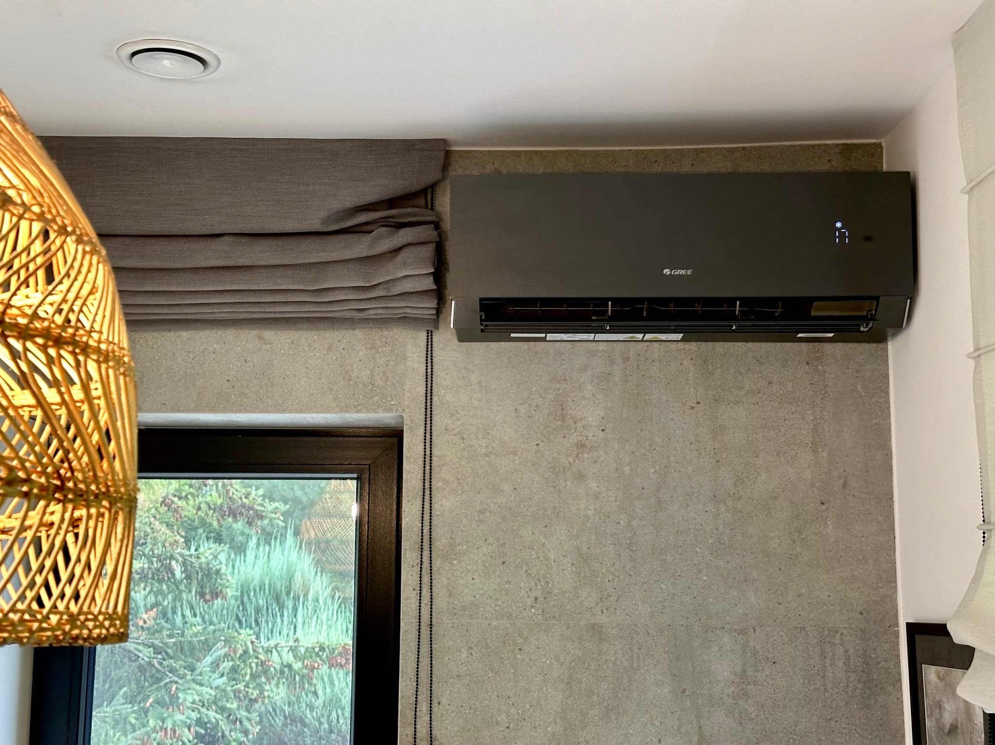 Szary klimatyzator marki Gree zamontowany na betonowej ścianie w nowoczesnym wnętrzu, widoczne rolety rzymskie i lampa z rattanu.