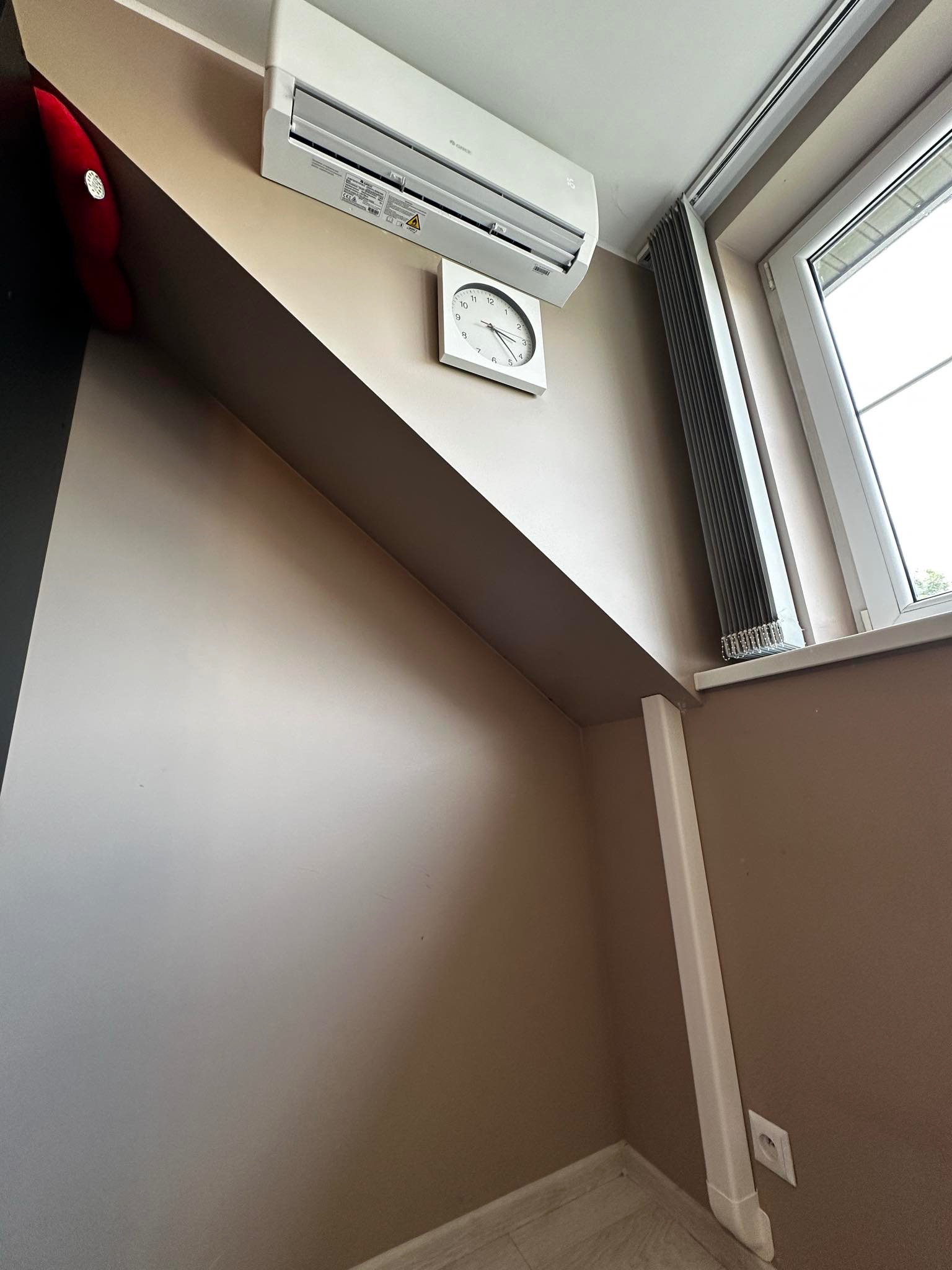 Biała klimatyzacja ścienna zamontowana w pokoju z beżowymi ścianami, obok okna z szarymi roletami i białym zegarem na ścianie.