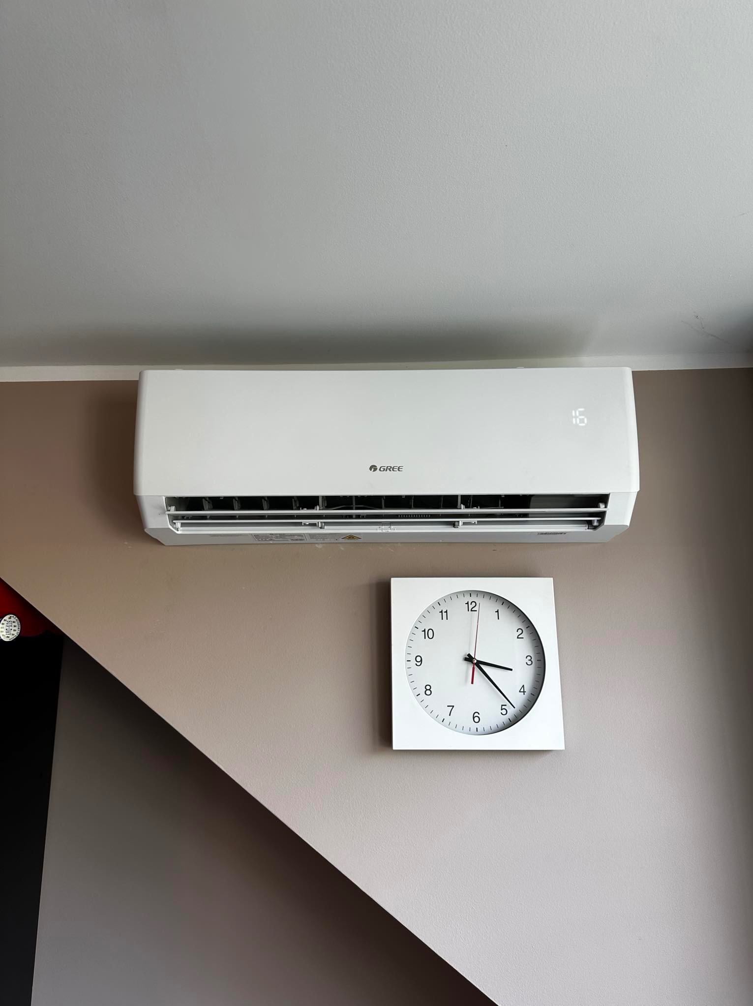 Biała klimatyzacja marki Gree zamontowana na ścianie w pomieszczeniu, obok biały zegar wskazujący godzinę 3:23, widoczny wyświetlacz z temperaturą 16 stopni.