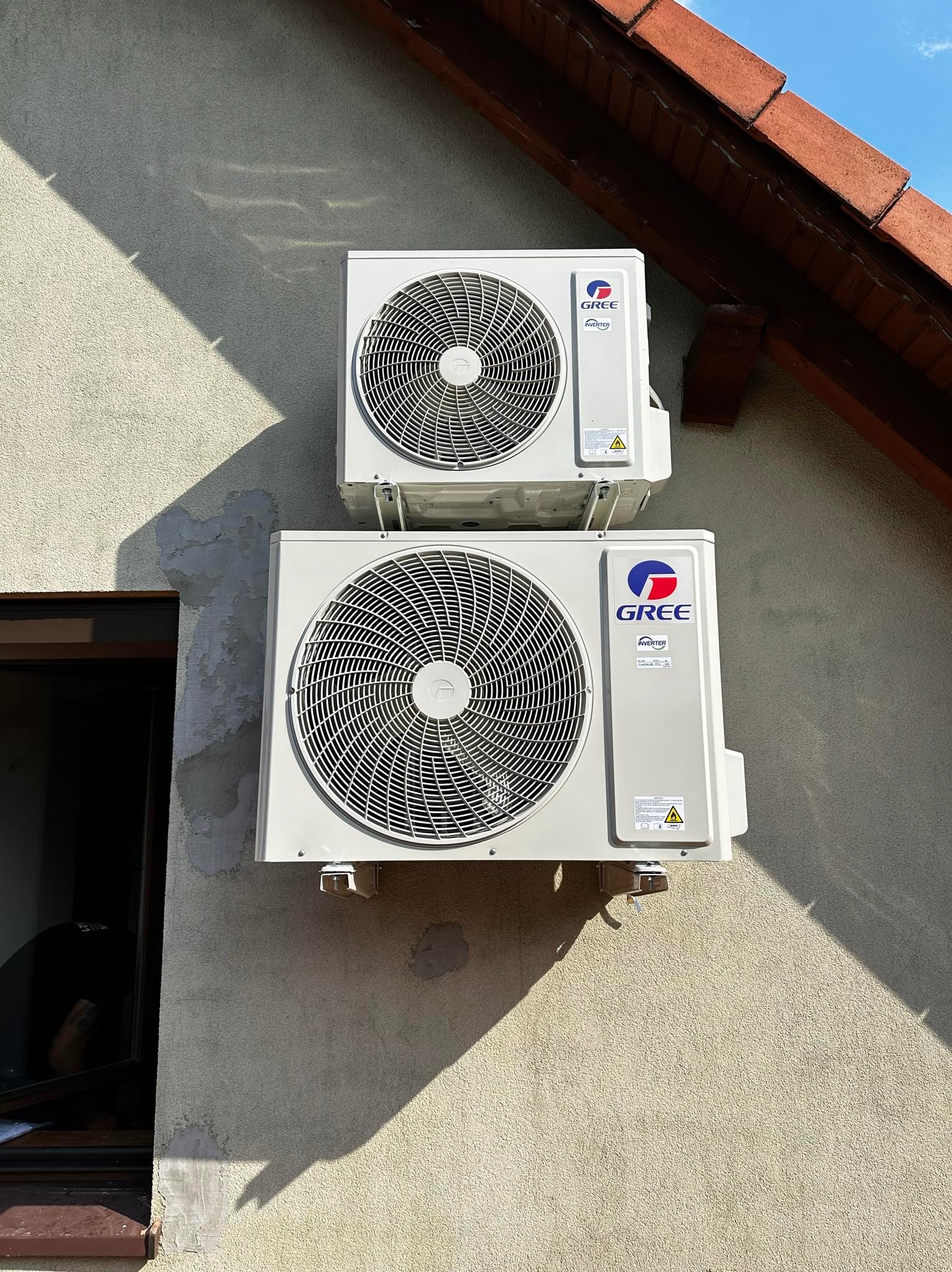 Dwa klimatyzatory marki Gree zamontowane jeden nad drugim na elewacji budynku, widoczne okno i fragment dachu.
