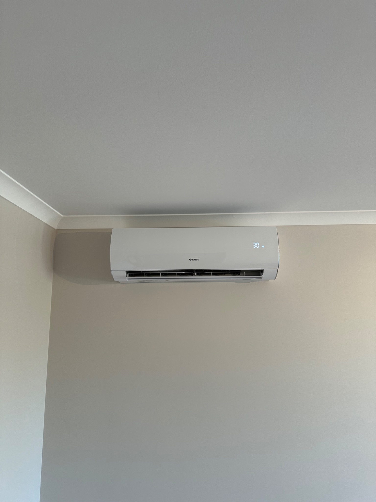 Biała jednostka klimatyzacji marki Gree zamontowana na beżowej ścianie w rogu pokoju, wyświetlająca temperaturę 30 stopni.