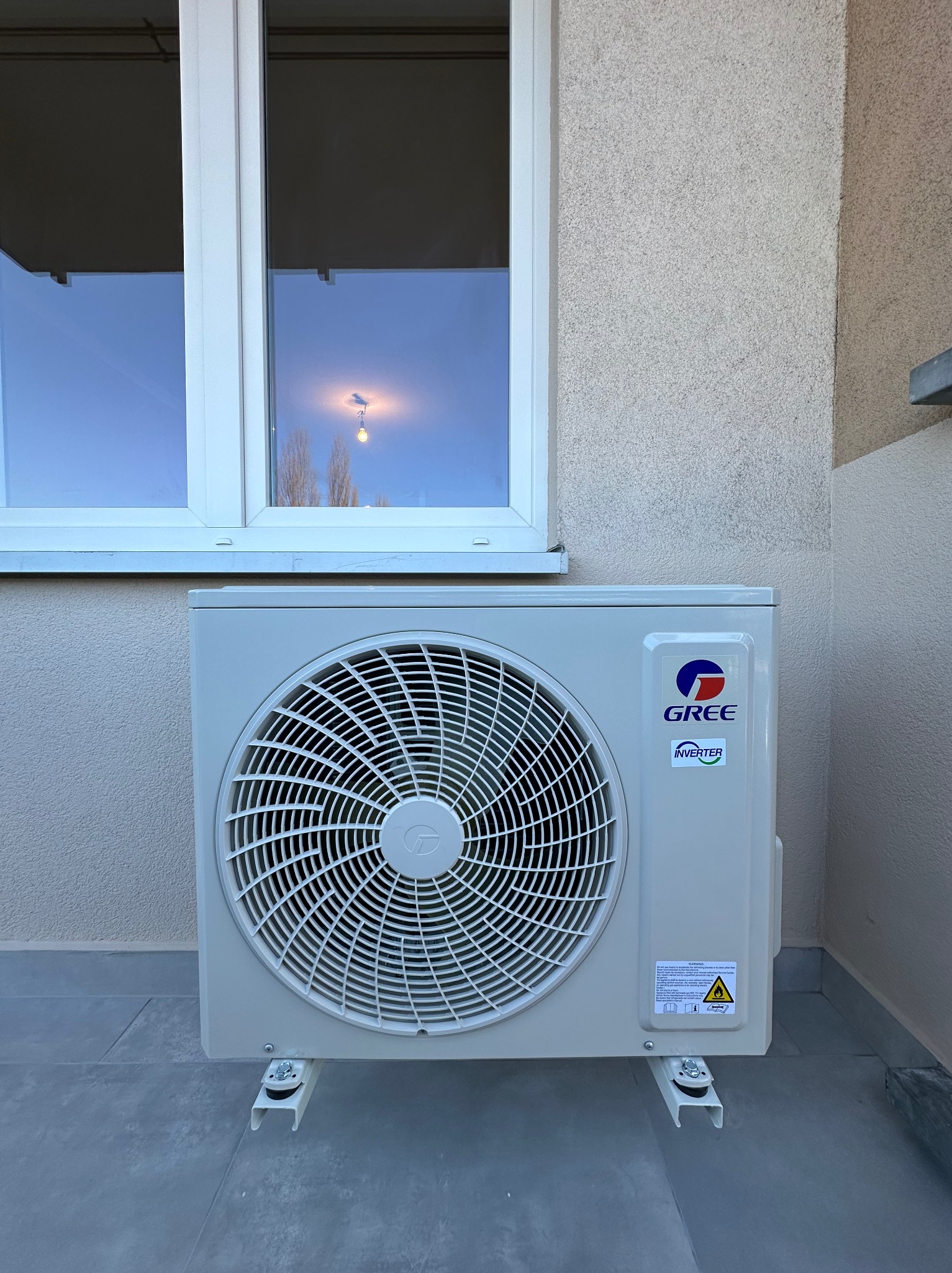 Zewnętrzna jednostka klimatyzacji marki Gree, zamontowana na elewacji budynku z widocznym oknem, oświetlonym żarówką w odbiciu szyby.