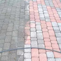 Filip Kopera - Mycie kostki brukowej: Widoczna różnica między brudną (szarą) a czystą (różowo-szarą) częścią kostki, z przewodem od myjki ciśnieniowej leżącym na kostce.