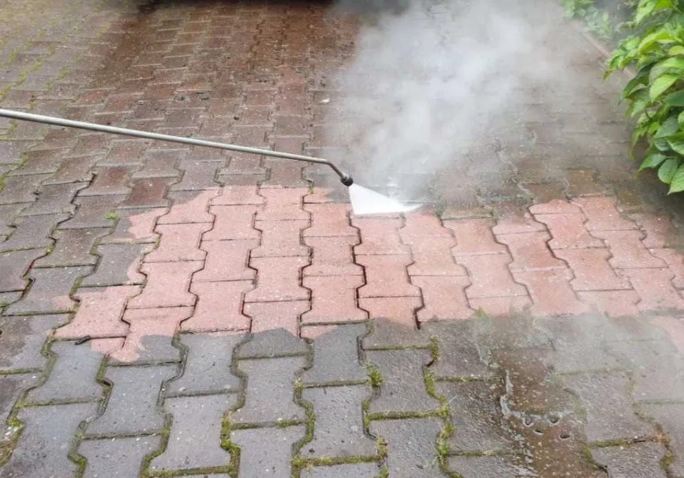 Częściowo wyczyszczona kostka brukowa w kolorze czerwono-szarym poddawana czyszczeniu pod ciśnieniem z widoczną lancą i strumieniem wody oraz parą wodną.