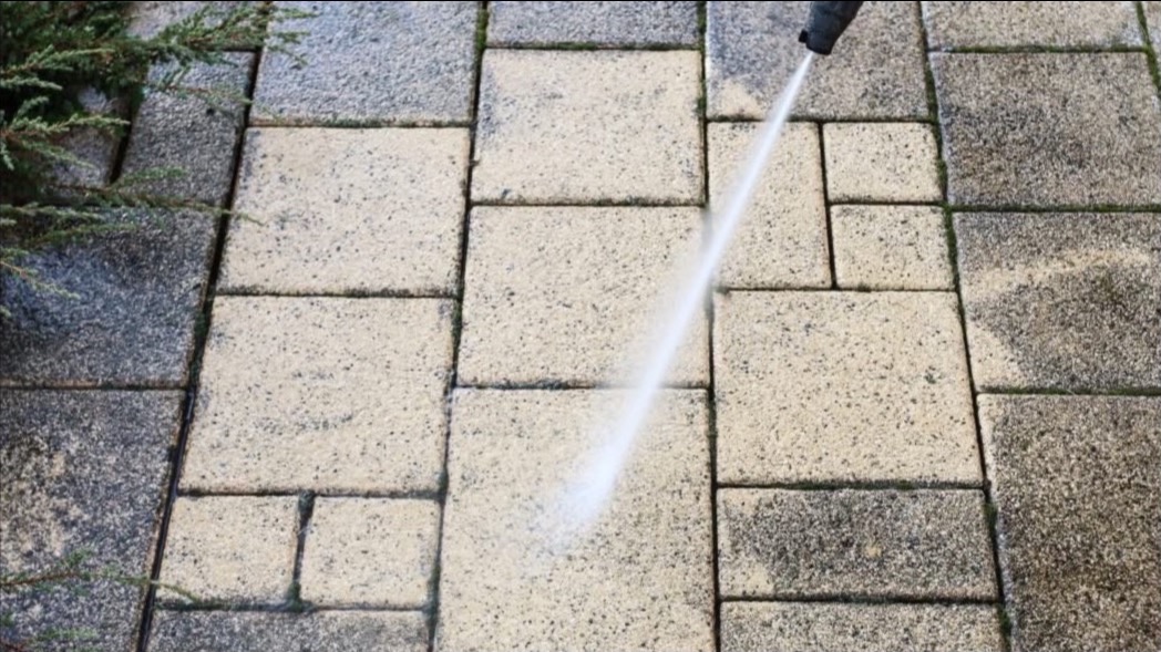 Mycie kostki brukowej z użyciem myjki ciśnieniowej, widoczna różnica między czystą a brudną powierzchnią kostki. Po lewej stronie widoczna roślinność.