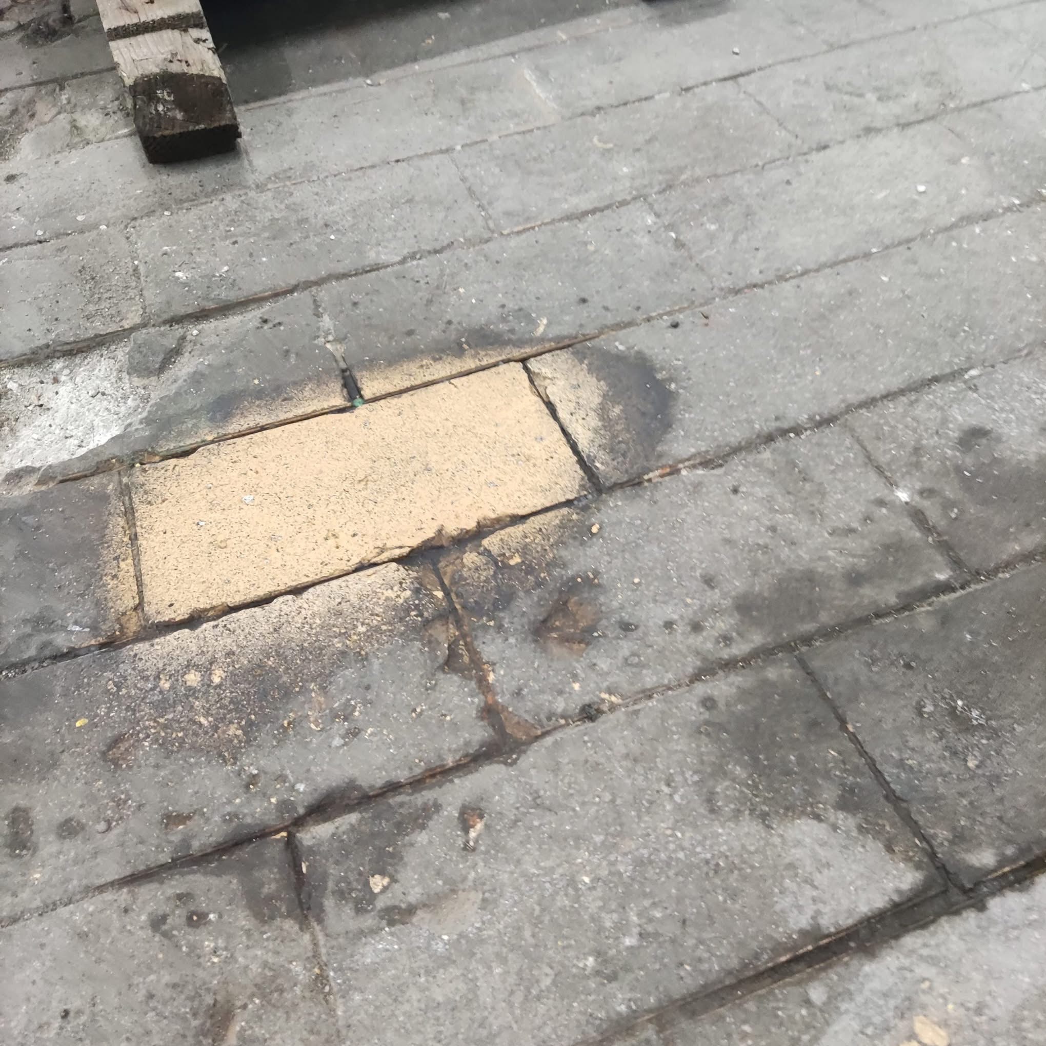 Fragment szarej kostki brukowej z pojedynczą, jasną kostką i zabrudzeniami, widoczne ślady wilgoci.