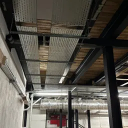 Sufit z instalacjami: koryta kablowe z perforowanej blachy, rury wentylacyjne w izolacji, konstrukcja stalowa. Widok z dołu na sufit w trakcie prac instalacyjnych.