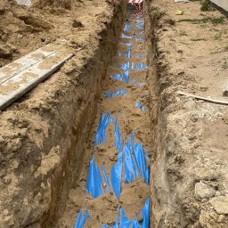 ElektroSync - Radosław Niżyński - Wykop z niebieską folią zabezpieczającą, prawdopodobnie pod instalację elektryczną. Widoczne materiały budowlane i domy w tle. Ujęcie z góry.