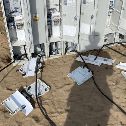 ElektroSync - Radosław Niżyński - Otwarta skrzynka elektryczna Inco-Bex 16/18 z widocznymi kablami i elementami instalacji na piaszczystym podłożu. Widoczne zabezpieczenia i skrzynki z numeracją.