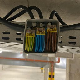 ElektroSync - Złącze elektryczne WAGO z przewodami w puszce instalacyjnej, zamontowane na metalowej szynie w garażu podziemnym. Widoczne kable w kolorach: zielono-żółtym, niebieskim i brązowym.