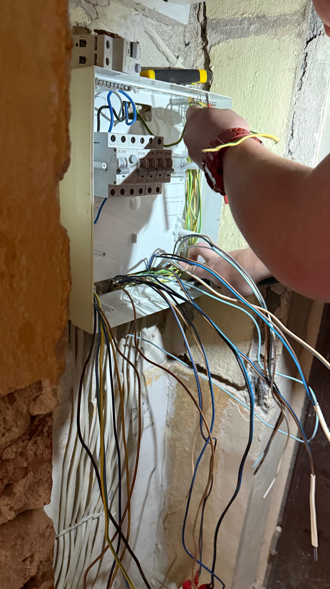 Elektryk łączy przewody w skrzynce rozdzielczej Eaton. Widoczne kable w różnych kolorach oraz narzędzia. Praca w trakcie wykonywania, surowe ściany w tle.